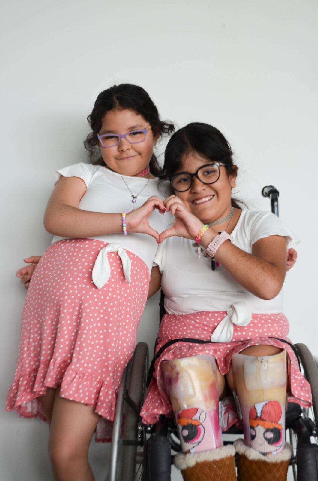 Dos niñas, una de ellas usuaria de silla de ruedas, sonríen abrazadas formando un corazón con sus manos