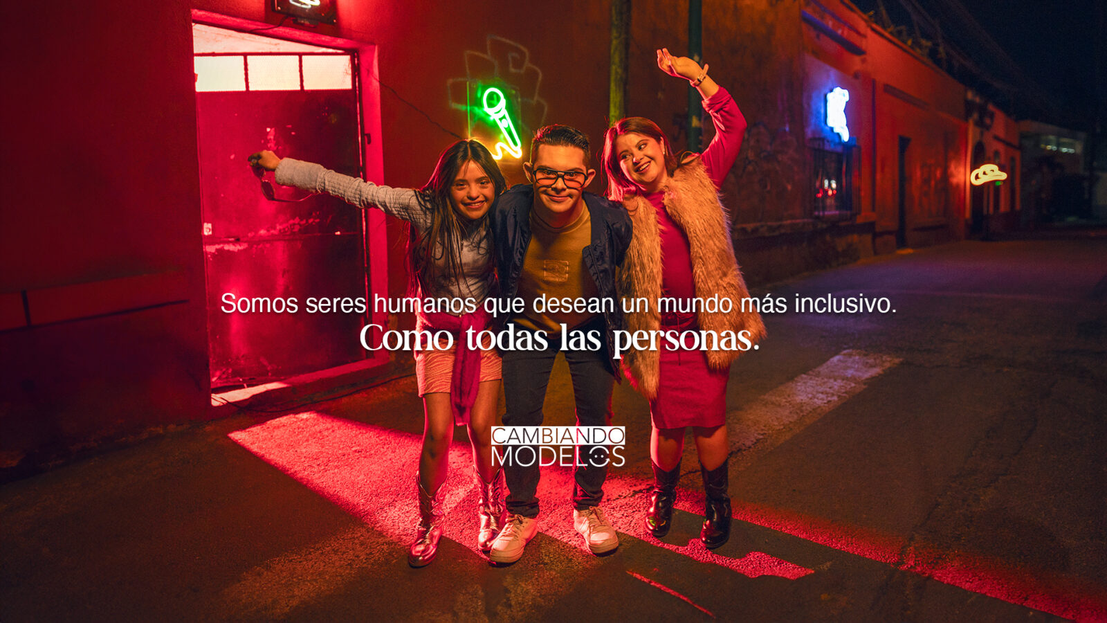 Tres jóvenes sonrientes, dos mujeres y un hombre con discapacidad, posan de noche frente a luces de neón con los brazos levantados celebrando la inclusión.